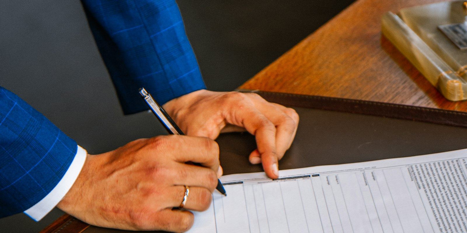 Mão de um homem com aliança assinando documentos em uma mesa